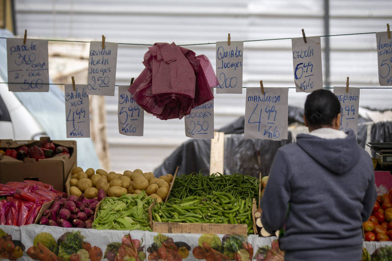 A imagem mostra uma barraca de feira com várias caixas de frutas e legumes. Há uma pessoa de costas， vestindo um moletom cinza， observando os produtos. Ao fundo， estão pendurados vários cartazes com preços e nomes de produtos， e uma folha de papel vermelha está pendurada no centro.