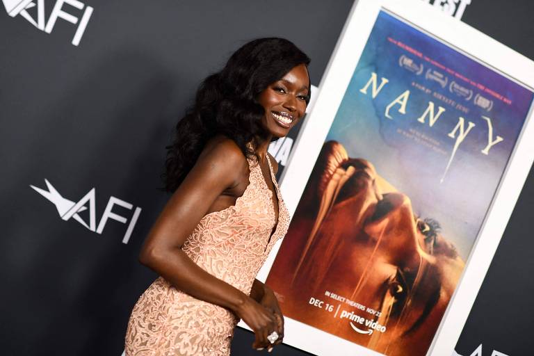 A atriz senegalesa, Anna Diop, durante a estreia de "Nanny", em 2022. Diop é uma mulher negra retinta e está usando um vestido de festa na cor rosa. O cabelo dela está liso e ela sorri ao lado do cartaz de divulgação do filme.