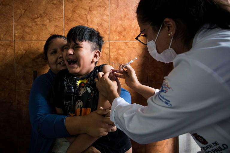 Imagem mostra mulher de jaleco branco, cabelos presos, óculos e máscara aplicando uma vacina num garotinho que está no colo de sua mãe, que encontra-se sentada; a criança está chorando porque não queria tomar a vacina