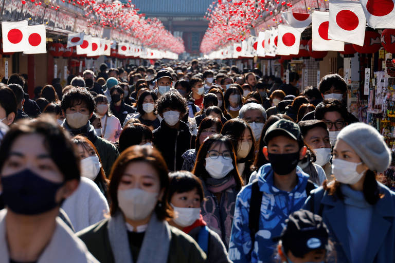 Pessoas, maioria asiática, de máscara, caminham em via com laterais cheias de bandeirinhasas do Japão