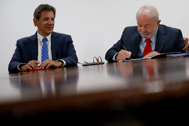 De terno escuro e gravata azul, o ministro da fazenda, um homem branco de cabelos castanhos não muito curtos e rosto barbeado, olha para o lado direito da imagem, onde aparece o presidente Lula, de cabelos brancos e barba branca, terno escuro e gravata vermelha. Ambos estão sentados a uma mesa que corta a imagem ao meio. Lula assina um papel com a mão direita, enquanto a esquerda firma  afolha sobre a mesa. Ao fundo, a parede é branca.; Entre os dois homens, sobre a mesa, está apoiado um par de óculos