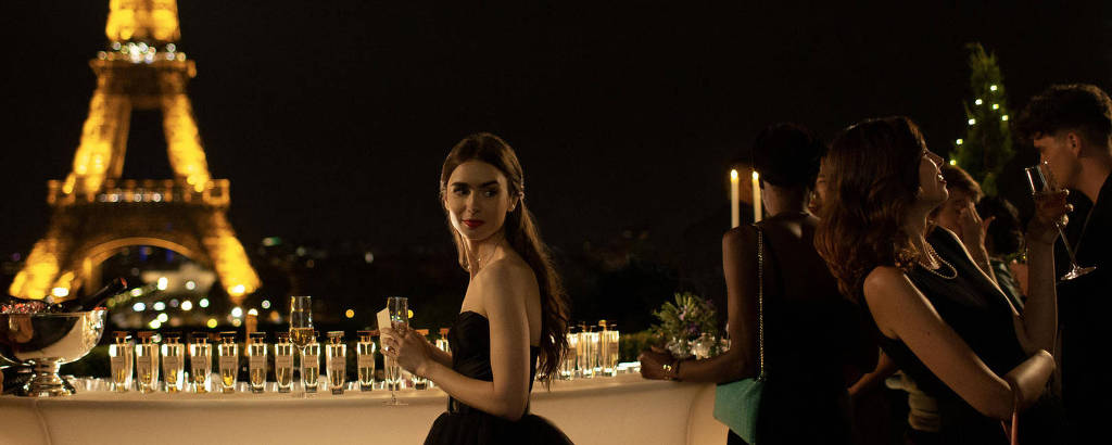 À noite, diante da Torrre Eiffel, mulher com um vestido preto segura uma taça de espumante e olha para outras pessoas, que conversam