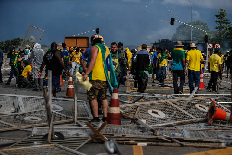 Grupo de pessoas com roupas verdes e amarelas e bandeiras brasileiras andam por uma grande praça pisando em destroços de barreiras de segurança  