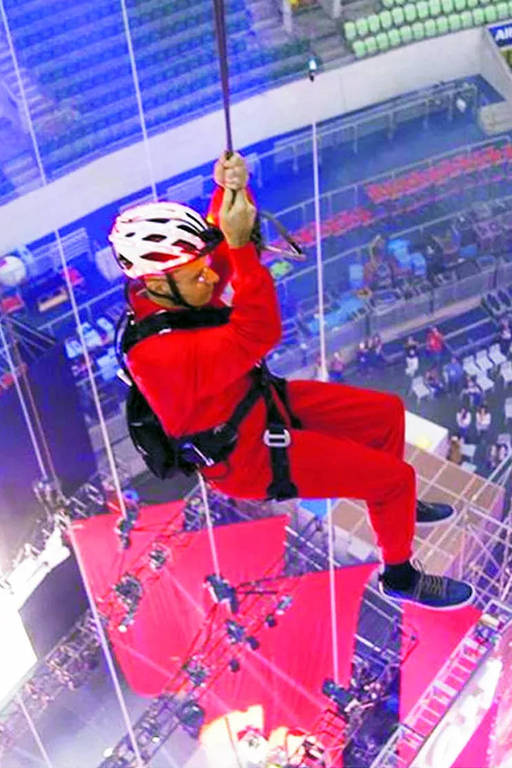 Homem de capacete branco e roupa vermelha segura em corda na qual está pendurado; ele [e visto do alto, com arquibancadas ao fundo