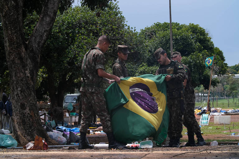 Quatro soldados fardados seguram bandeira do Brasil recolhida de acampamento, ao fundo árvores e na grama, objetos remanescentes do acampamento bolsonarista, céu azul
