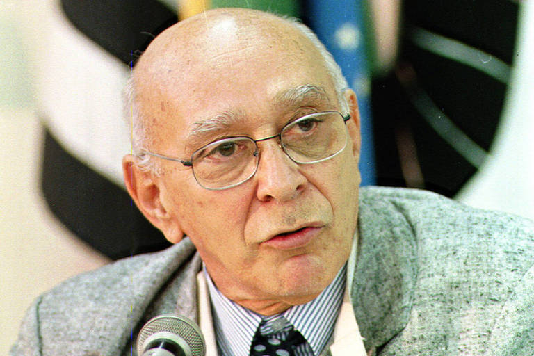 Um homem idoso com cabelo calvo e óculos está falando em um microfone. Ele usa um paletó cinza e uma gravata listrada. Ao fundo, há bandeiras e um ambiente de conferência.