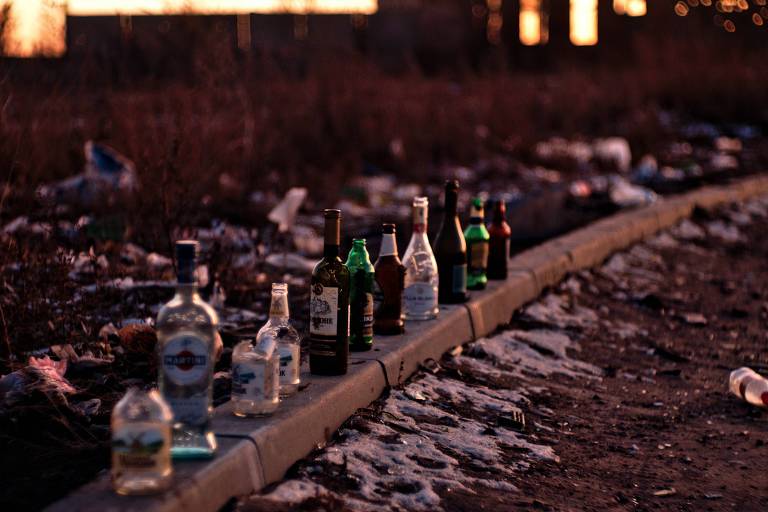 Foto com vista de parte da rua e da calçada mostra garrafas de bebida enfileiradas