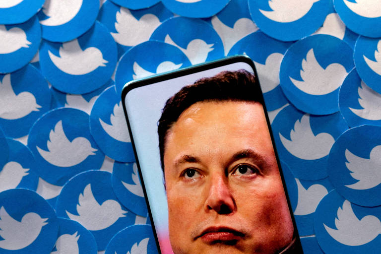 Elon Musk é retratado na tela de um smarphone. Ao fundo, há várias impressões do logo do Twitter, plataforma comprada pelo bilionário em 2022.