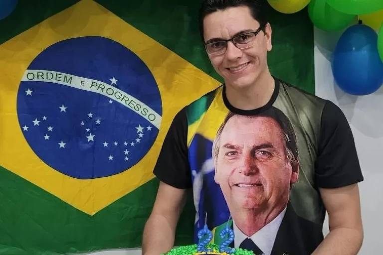 Homem usando camiseta estampada com a fotografia de Bolsonaro posa para foto. Ao fundo, há uma bandeira do Brasil