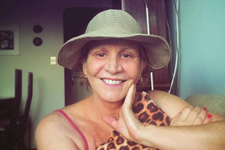 Rosângela Alamini, branca, sorri para foto, usa chapéu e vestido de praia com uma alça. Apoia mão esquerda na bocheca esquerda e mão direita no braço esquerdo