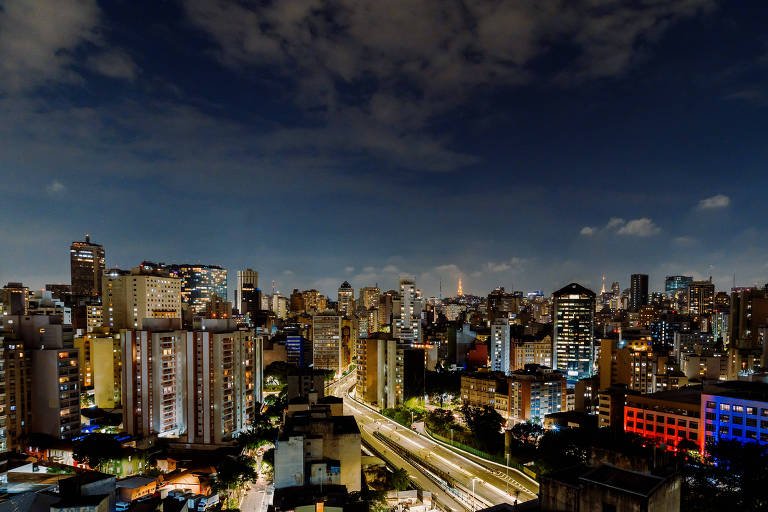 Imagem mostra diversos prédios no horizonte, à noite, com as luzes da cidade clareando o céu