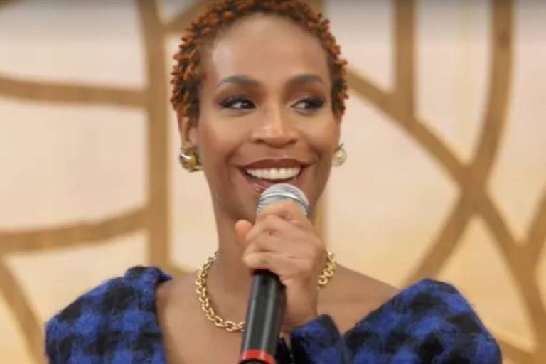 Em foto colorida, mulher sorri ao segurar um microfone para cantar em um programa de TV