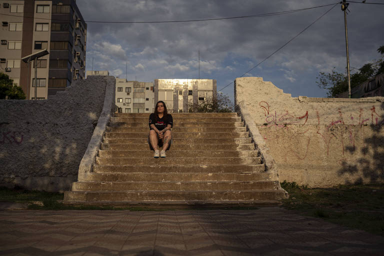Em uma escadaria de concreto ao ar livre, uma mulher branca de saia jeans e camiseta preta está sentada ao ar livre.