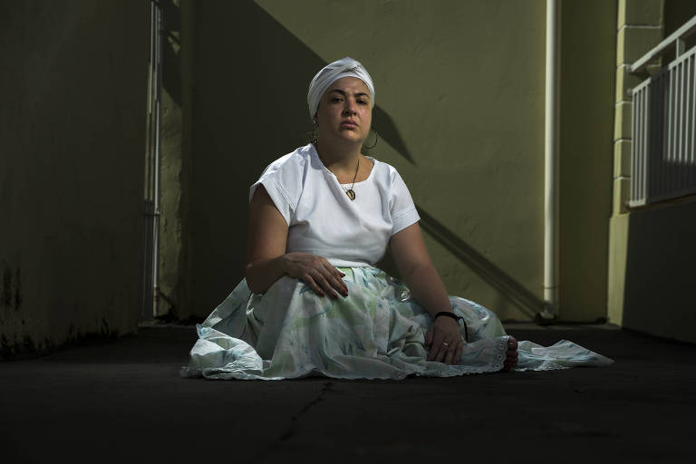 Juliana Arcanjo Ferreira está séria, sentada no chão e usa saia longa e turbante