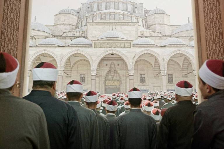 Homens em mesquita são vistos de costas para a foto, todo com chapéus vermelhos e vestes iguais