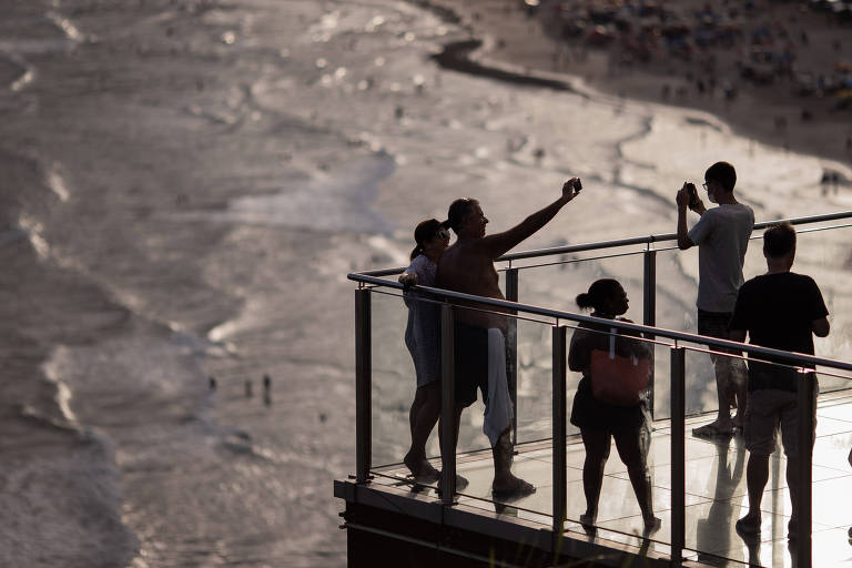 foto mostra silhueta dos turistas, que tiram fotos e selfies e, ao fundo, o mar