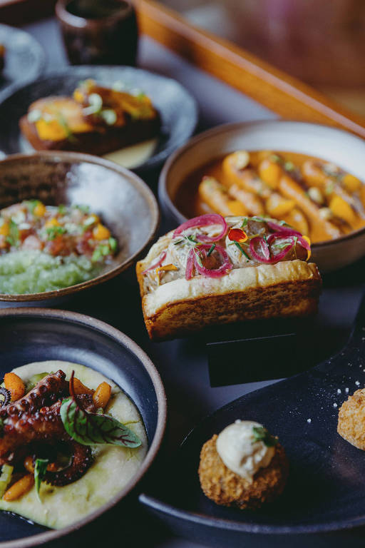 A foto mostra pratos variados sobre um carrinho que circula pelo restaurante Animus. Há polvo, sanduíche de camarão, bolinhos fritos e cenouras na brasa 