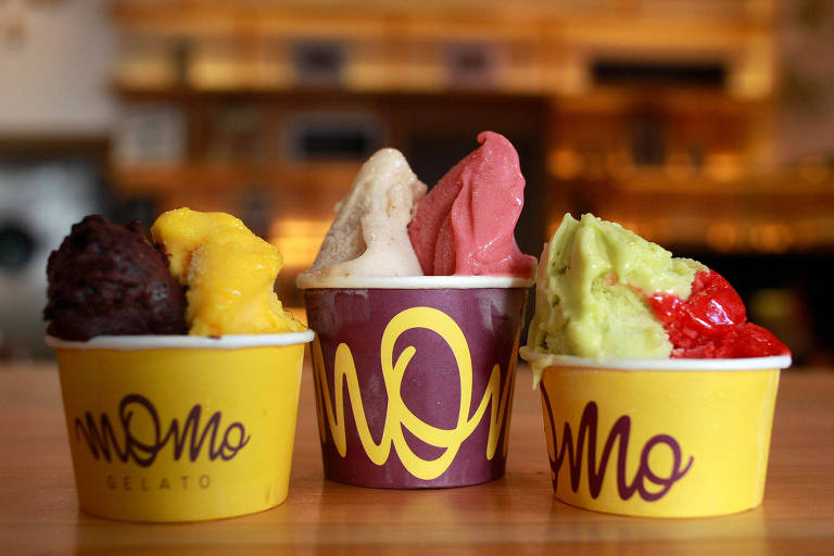 A foto mostra três copinhos de papel nas cores amarelo e roxo, com gelatos de diversos sabores