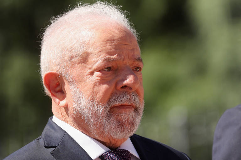 A foto mostra o rosto de Lula, um homem branco, de barba e cabelos brancos, de terno. Ele aparece em close, olhando para a frente