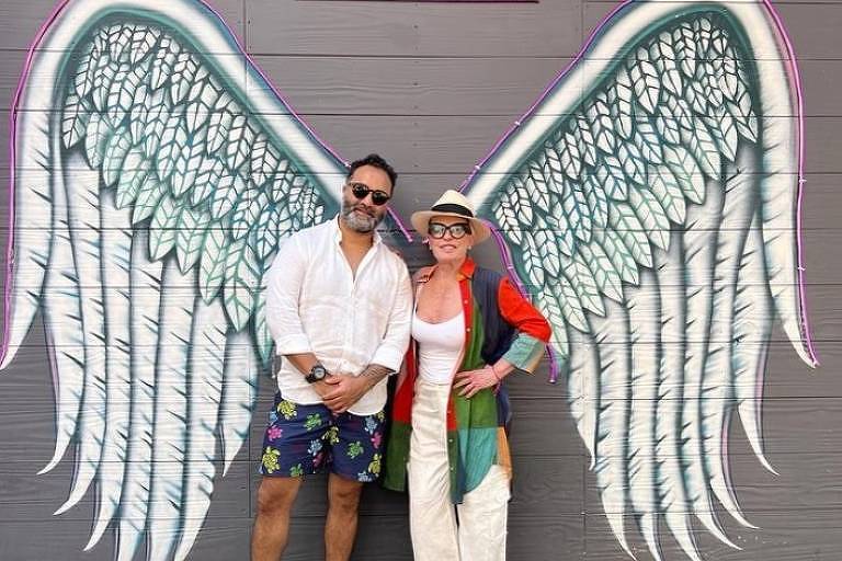 Em foto colorida, um homem e uma mulher posam juntos em um muro com asas de anjo