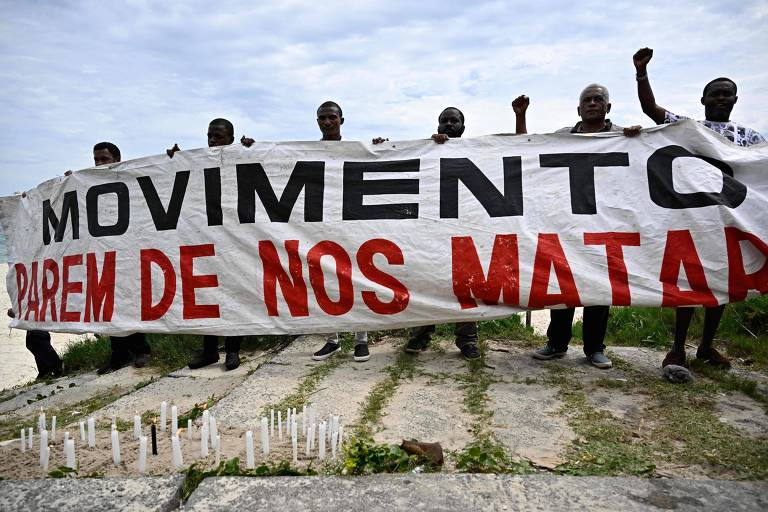 Imagem colorida mostra homens negros segurando uma faixa branca escrito "Movimento - parem de nos matar" nas cores preta e vermelha. À frente há velas acesas em homenagem ao congolês morto no Rio.