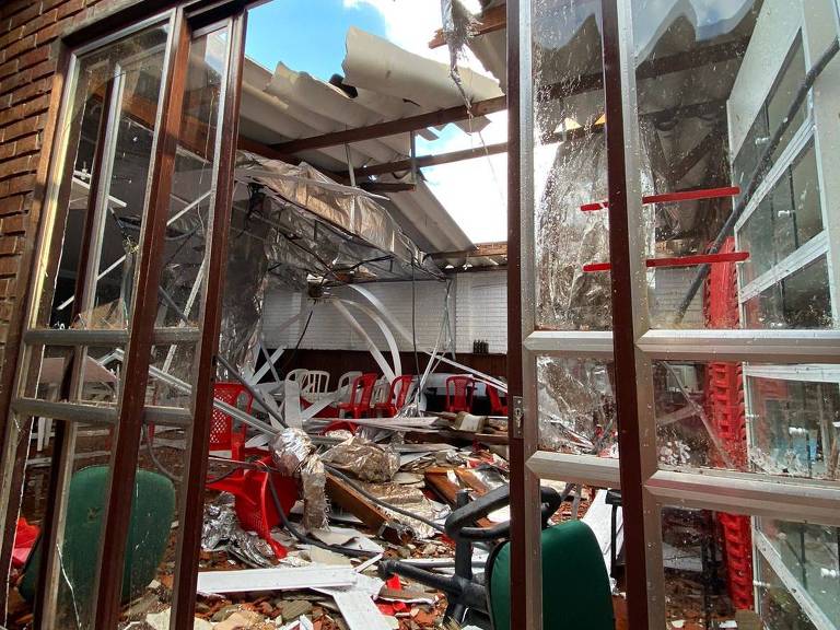 Porta aberta mostra sala com cadeiras viradas, destroços de pedra, madeira e telhado que sobraram do centro espírita atingido pela parede que desabou do 20° andar de um prédio ao lado