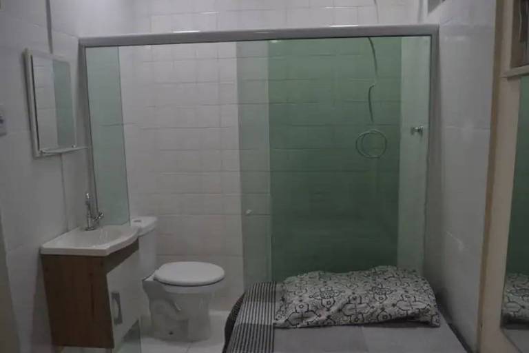 Banheiro com azulejos brancos, pia e espelho, ao lado de uma cama com lençol cinza, sobre o qual está um travesseiro. Ao fundo, há um box de vidro, com uma privada e um chuveiro.