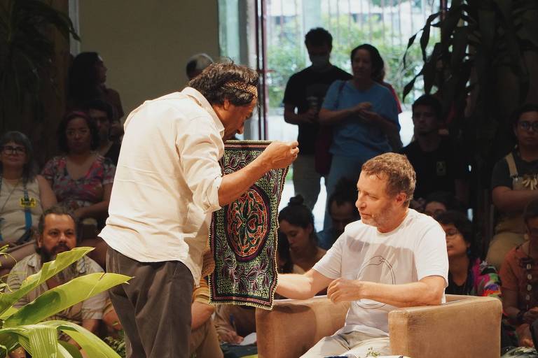 Homem de pé mostra pedaço de tecido colorido para outro homem, sentado em uma poltrona. Os dois estão cercados de pessoas sentadas em sala. 