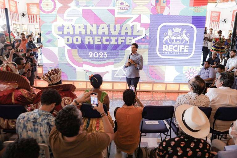 Prefeito do Recife, João Campos ao centro do palco e à frente de um telão com os dizeres "Carnaval do Recife 2023" falando para o público durante anúncio da programação da festa.