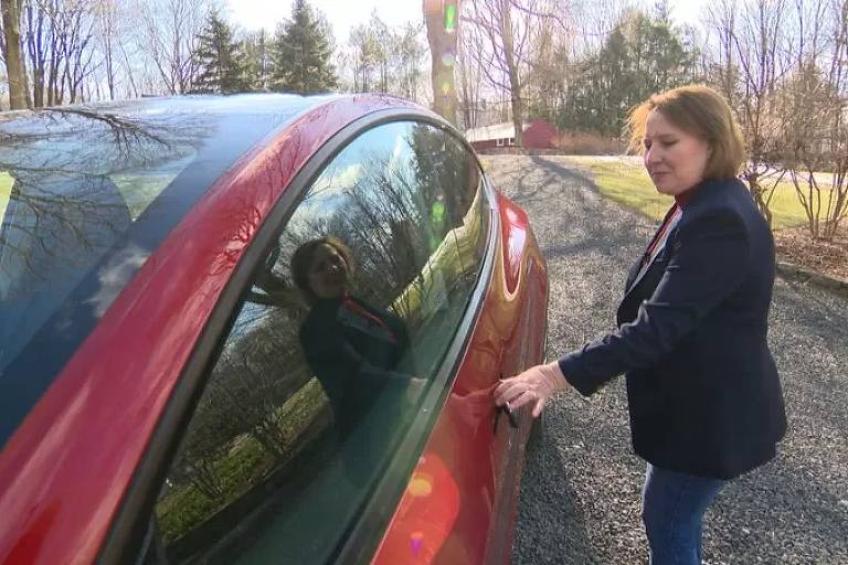 Uma mulher de 55 anos, vestida de jeans e agasalho, abre a porta de seu veículo Tesla vermelho, à sua esquerda. O cenário de inverno é tomado por pinheiros e árvores caducifólias