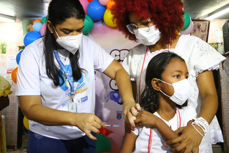 criança de máscara é vacinada por profissional de saúde de máscara com mulher atrás de máscara; ao fundo, bexigas coloridas