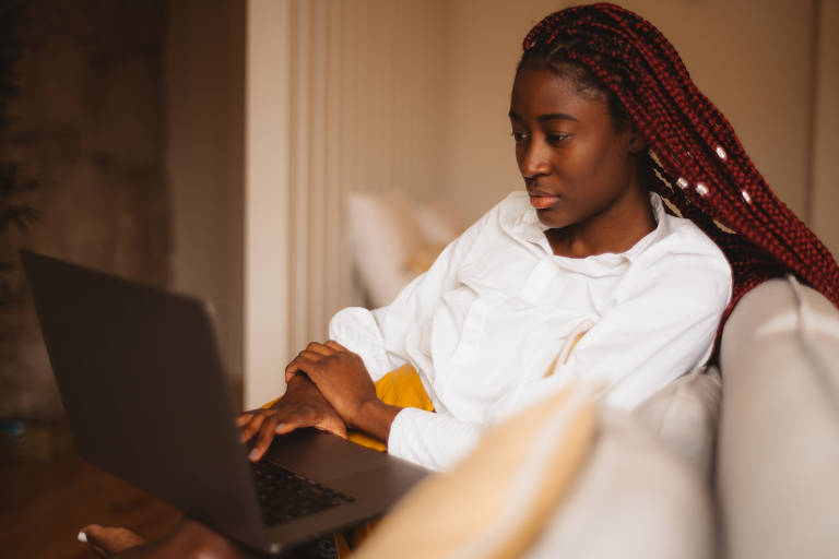 Um retrato de uma bela jovem aluna negra com tranças de caixa de cor rosa estudando em seu laptop enquanto está sentada no sofá bege em sua sala de estar, com um fundo desfocado
