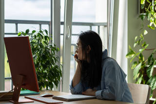 Thoughtful young Asian woman sits at desk with computer trying to come up with resume for job search