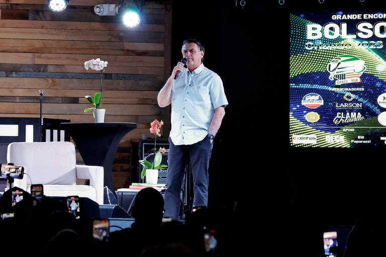 De camisa azul clara e jeans sobre o palco e diante de um telão, Bolsonaro, um homem branco de cabelos lisos e castanhos, discursa para apoiadores