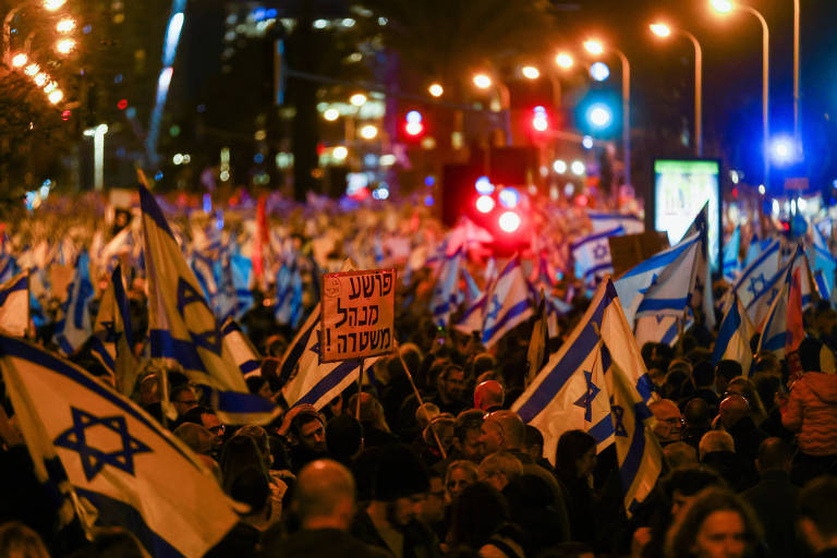 Milhares de pessoas são vistas em uma avenida cercada de prédios balançando bandeiras de Israel, que tem uma estrela azul no centro