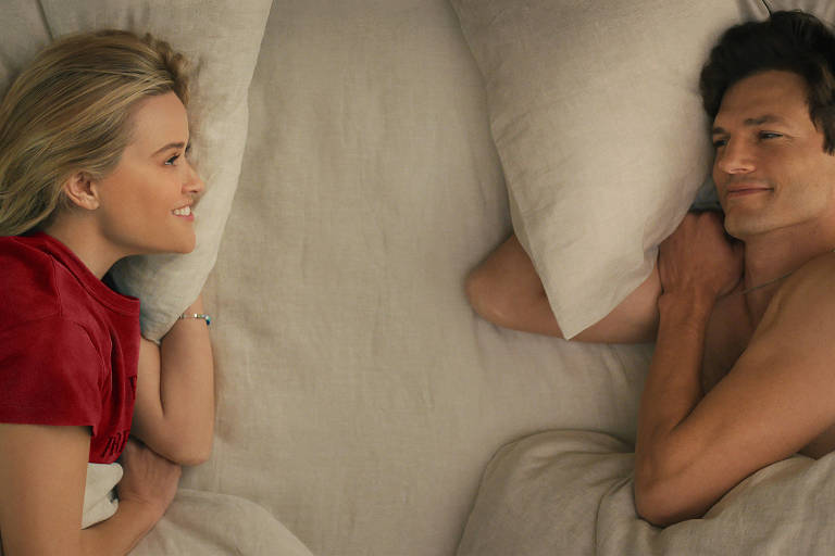 Reese Witherspoon e Ashton Kutcher como Debbie e Peter em "Na Sua Casa ou na Minha?".O casal está deitado em uma cama com lençóis e travesseiros brancos. Eles se olham e riem. Debbie veste uma camiseta vermelha e Peter está sem camisa. Ela é branca, loira e tem os olhos verdes. Ele é branco, moreno e com olhos escuros.