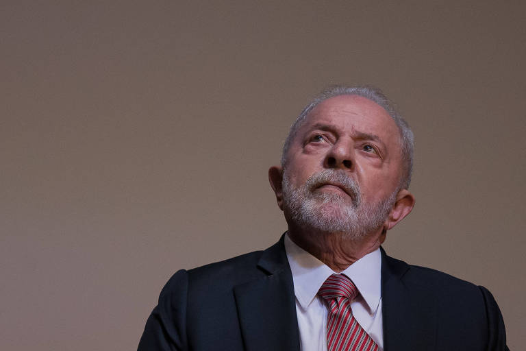 Lula está com a cabeça erguida, olhando para cima. Ele usa um terno preto com gravata vermelha.