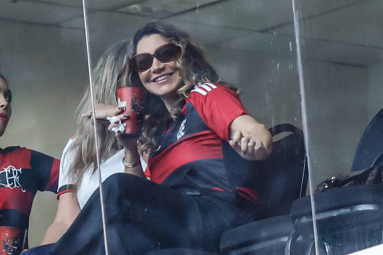 Em foto colorida, mulher de camisa rubro-negra aparece sentanda em uma tribuna de honra de um estádio de futebol