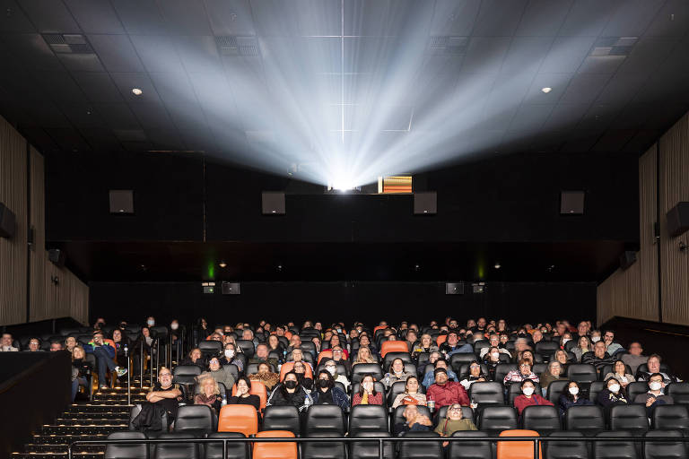 Imagem mostra poltronas ocupadas por pessoas em uma sala de cinema. Em cima delas, é possível ver a luz que projeta o filme na tela.