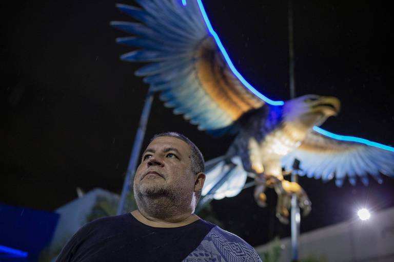 Homem branco olha imponente para cima; atrás dele, há uma enorme águia azul e branca iluminada