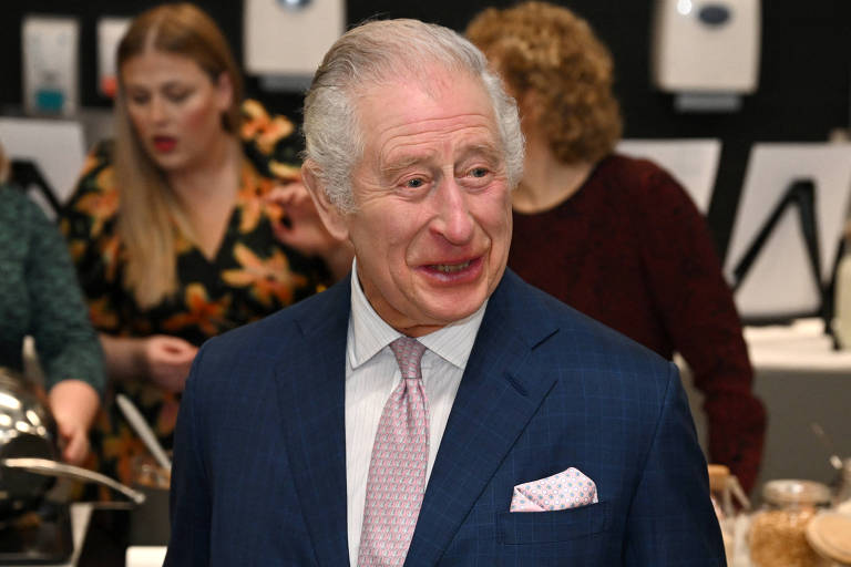 Rei Charles 3º sorri. Ele veste um terno azul marinho com gravata rosa. Ao redor dele, há outras pessoas.