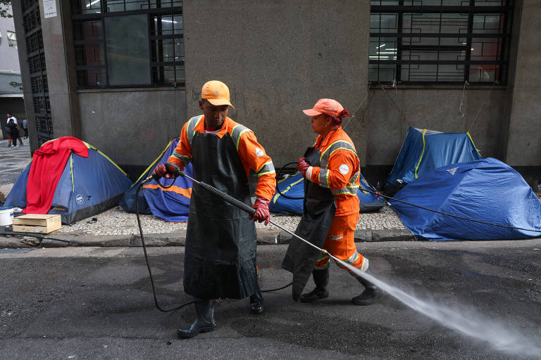 Duas pessoas com bonés e uniformes laranjas estão no centro da imagem, com barracas de lona azul ao fundo; uma das pessoas, que usa também um avental preto, limpa a rua com um jato d'água