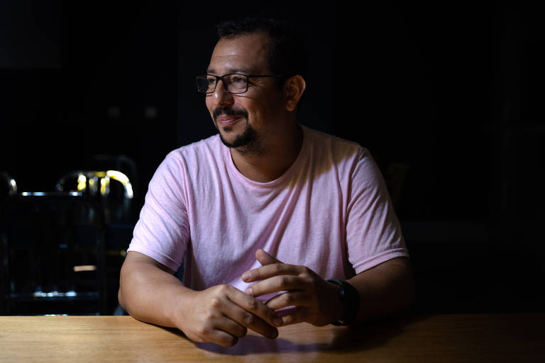 Retrato do professor Rodrigo Chagas, da Universidade Federal de Roraima. Ele está sentado, com as mãos sobre uma mesa de madeira, veste uma camiseta rosa e um relógio de pulso. Usa óculos e um cavanhaque