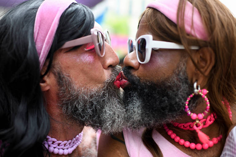 Fotografia colorida em primeiro plano mostra dois homens de perfil tocando os lábios em um beijo; eles têm barba e estão fansiados de mulher, com roupas cor-de-rosa
