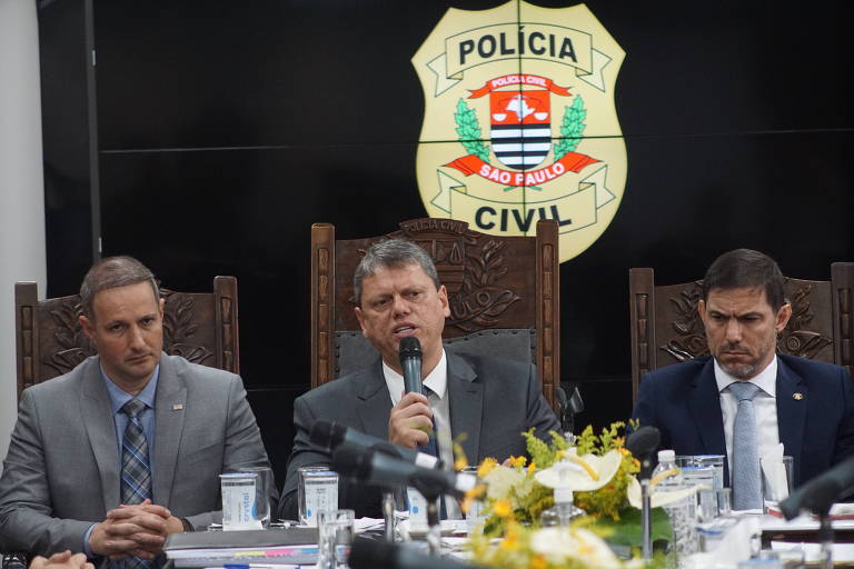 Da esquerda para a direita: Guilherme Derrite, Tarcísio de Freitas e Artur Dian. eles estão sentados atrás de mesa, e tarcisio discursa ao microfone