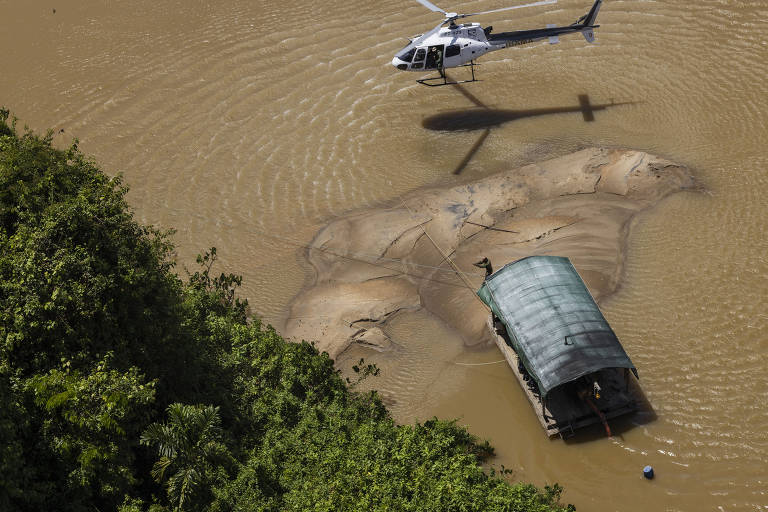 foto aérea mostra helicóptero sobrevoando o rio, onde há uma balsa de garimpeiros. Rio está lamacento