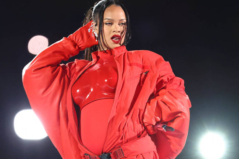 Rihanna de roupa vermelha e microfone na mão bota a mão no ouvido como quem diz "quero escutar vocês"