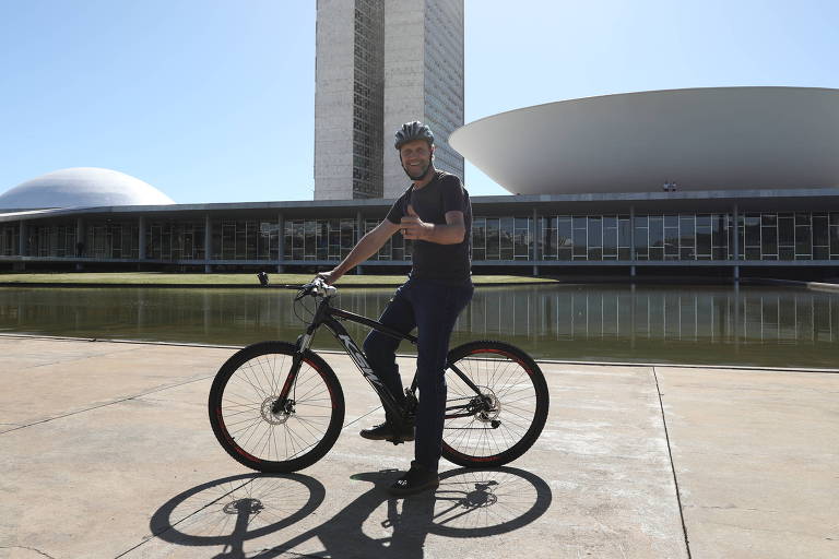 Vestindo capacete, calça e camiseta, homem faz sinal de positivo sobre bicicleta em frente ao prédio do Congresso Nacional