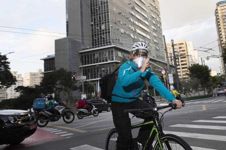 De máscara e jaqueta azul, Falzoni pedala em frente ao prédio da Câmara Municipal