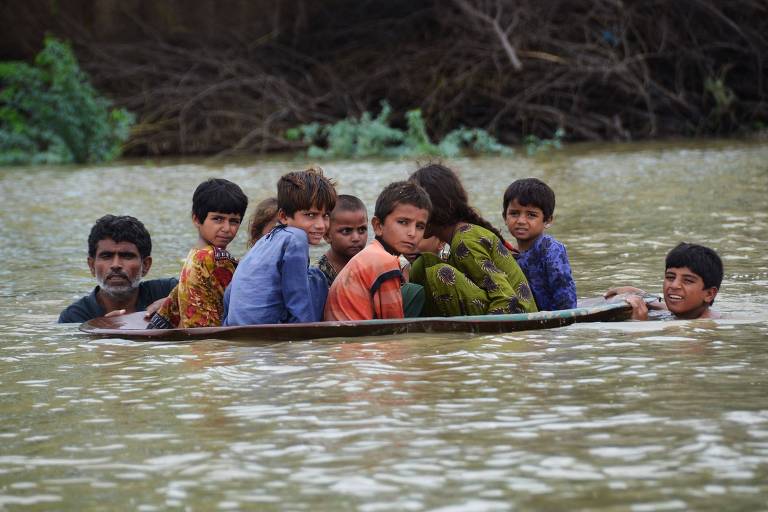 Grupo de crianças apertadas em um bote improvisado com dois homens as conduzindo com a água na altura do pescoço